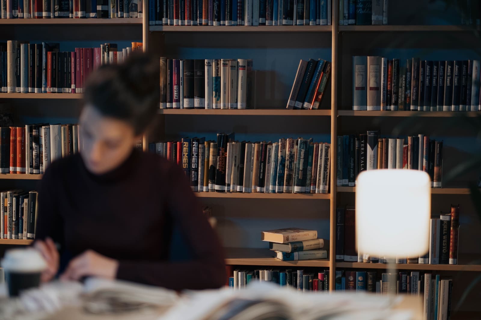 A student studying at a desk lit by a small lamp, in front of full bookshelves