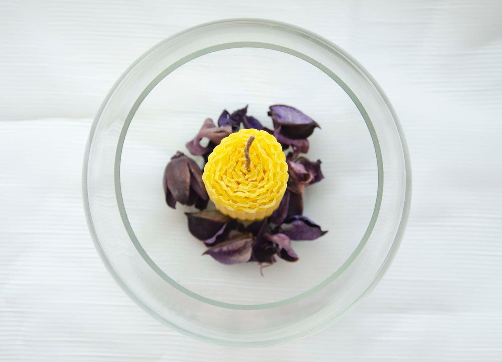 A rolled beeswax candle in a glass bowl of dried purple petals, seen from above
