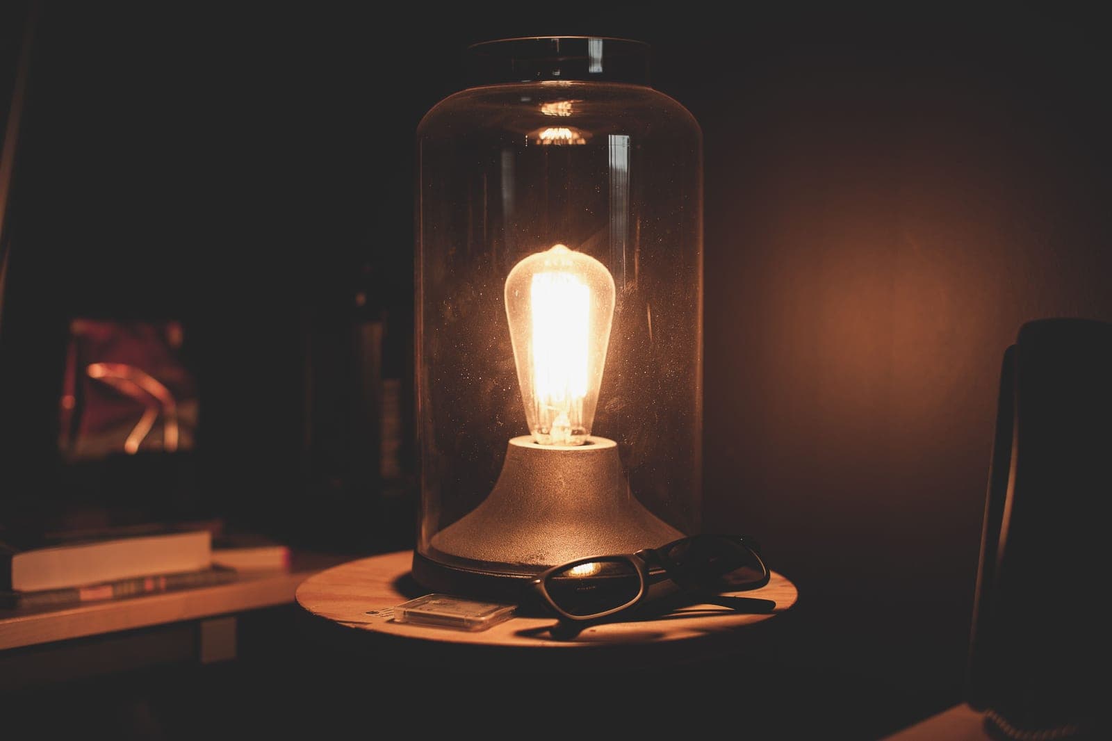 A warm table lamp glowing beside a pair of glasses in a dark room