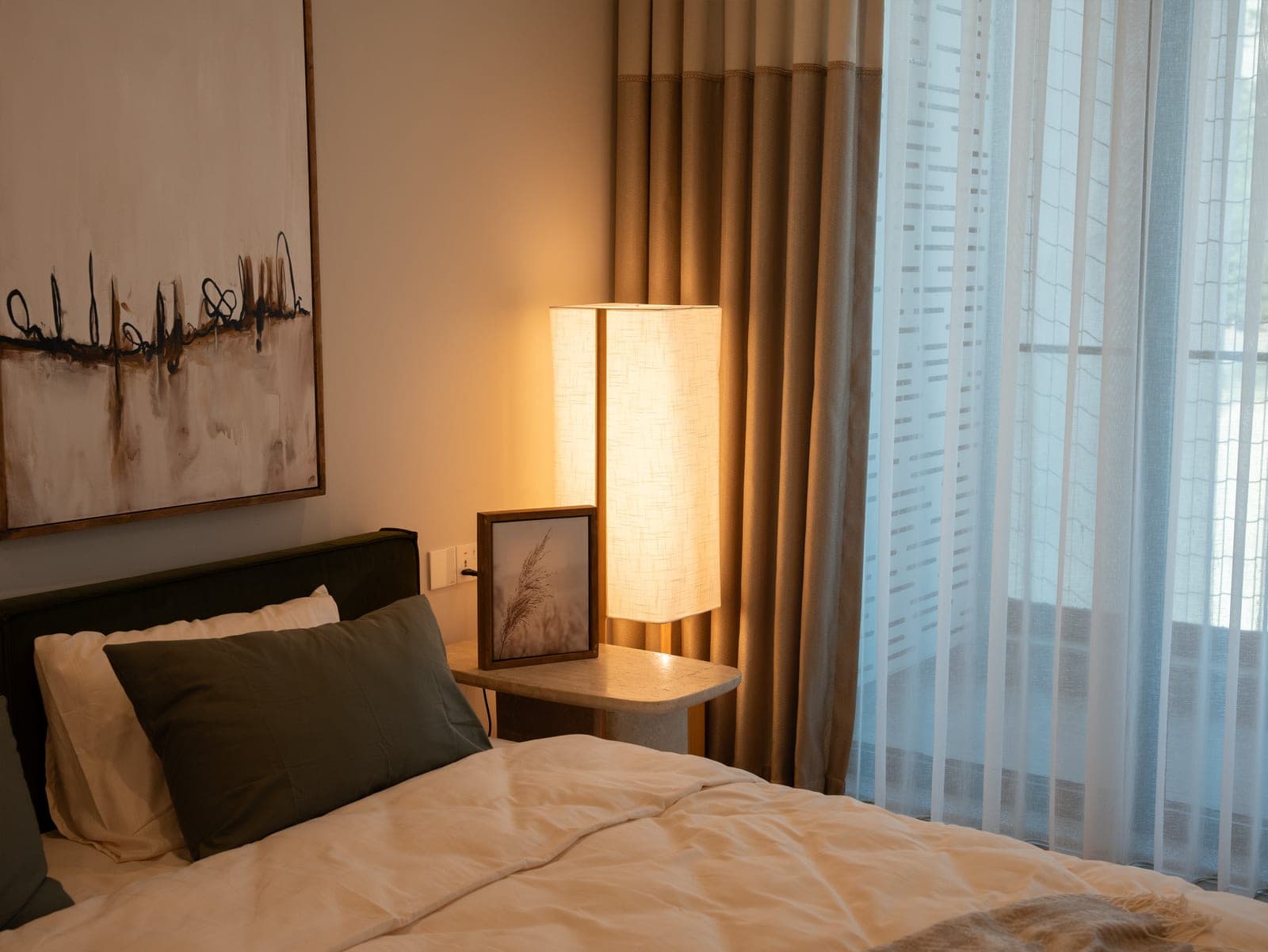 A quiet bedroom corner, a shaded lamp lit beside the bed and daylight through sheer curtains